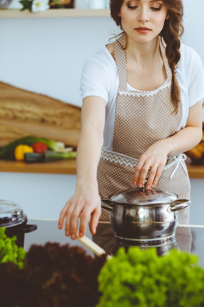 Giovane donna bruna che cucina zuppa in cucina, casalinga con un cucchiaio di legno in mano, concetto di cibo e salute.