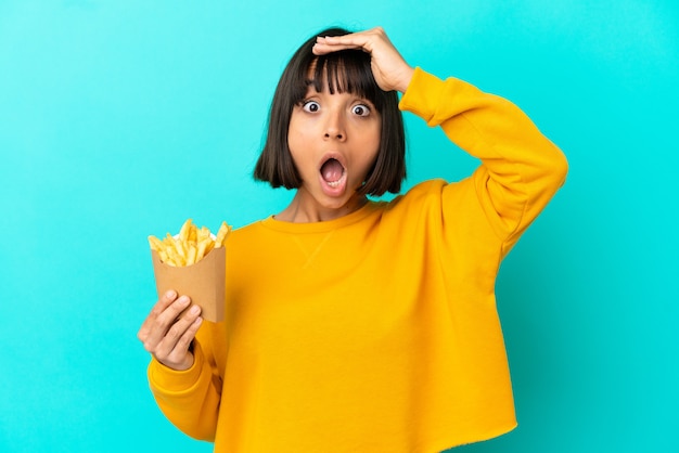 Giovane donna bruna che tiene patatine fritte su una superficie blu isolata che fa un gesto a sorpresa mentre guarda di lato