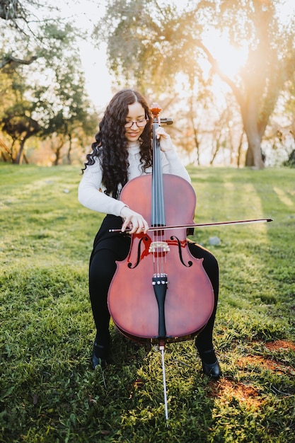 Giovane donna bruna con gli occhiali che suona il violoncello al tramonto nel parco, su un prato verde. Verticale