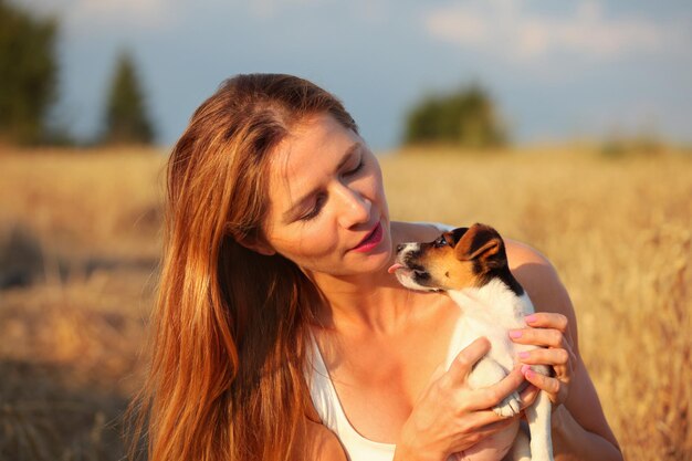 Giovane donna bruna con i capelli lunghi, con in mano un cucciolo di Jack Russell terrier che sta cercando di leccarla sul viso. Campo di grano illuminato dal sole pomeridiano sullo sfondo.