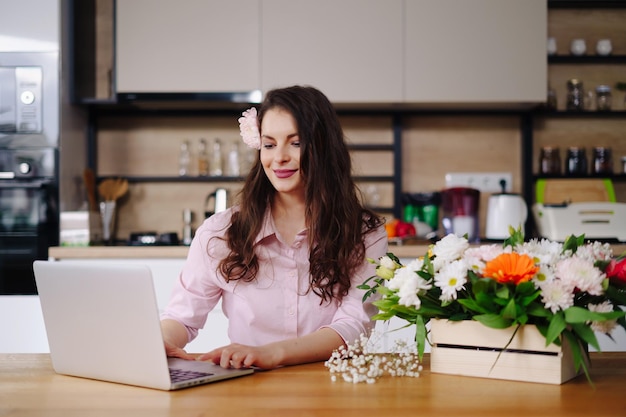 Giovane donna bruna con lunghi capelli ondulati che lavora al computer portatile con fiori sulla scrivania con cucina sullo sfondo Fiorista di talento che sviluppa vendite online e si prepara per l'officina