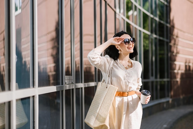 Giovane donna bruna con occhiali da sole e borsa che tiene caffè a piedi in città. Ritratto di stile di vita di donna