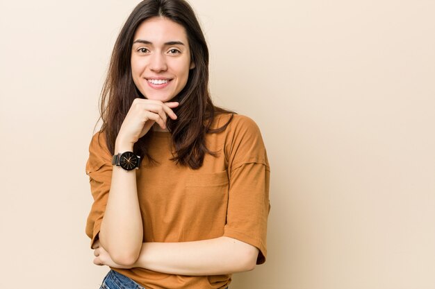 Giovane donna bruna contro un muro beige sorridendo felice e fiducioso, toccando il mento con la mano.