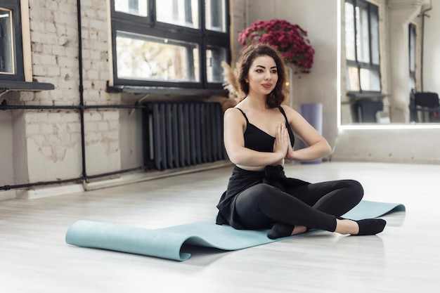 Giovane donna bruna in abiti sportivi si siede sul tappetino e medita nello studio di yoga