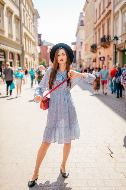 Giovane donna bruna in abito elegante e cappello in posa sulla strada della città