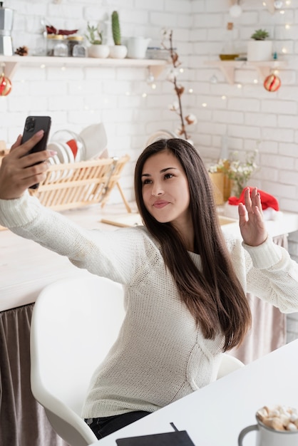 Giovane donna bruna in chat con gli amici utilizzando il suo telefono cellulare in cucina