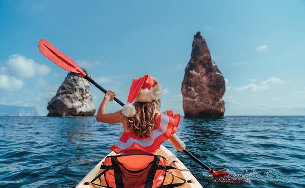 Giovane donna bruna in costume da bagno rosso e cappello da Babbo Natale che nuota in kayak intorno a rocce di basalto come dentro