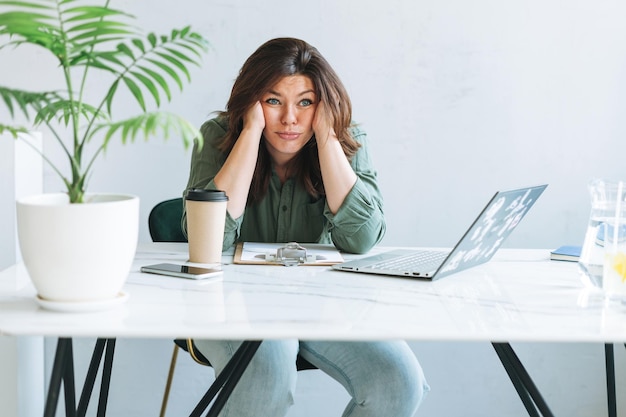 Giovane donna bruna infelice di pensiero più dimensioni che lavorano al computer portatile sul tavolo con pianta di casa nell'ufficio moderno e luminoso