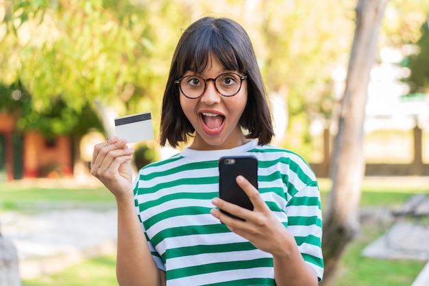 Giovane donna bruna nel parco che compra con il cellulare e tiene in mano una carta di credito con espressione sorpresa