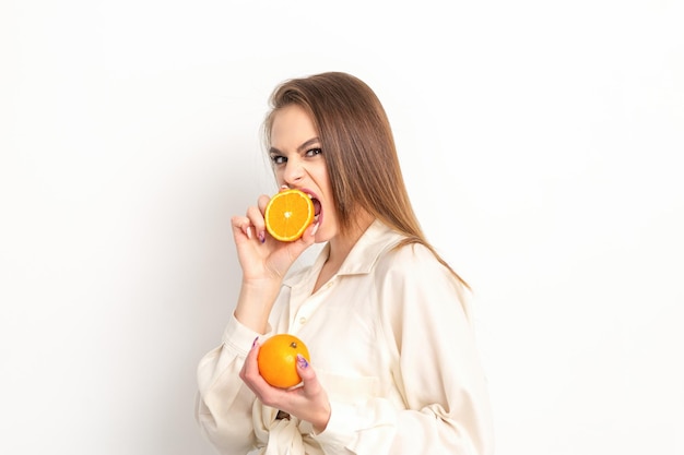 Giovane donna bruna piuttosto astuta caucasica morde una metà arancione e guardando la telecamera che indossa una camicia bianca su sfondo bianco