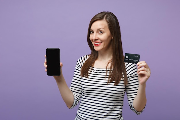 Giovane donna bruna ragazza in abiti casual a righe in posa isolata su sfondo viola viola ritratto in studio. Concetto di stile di vita delle persone. Mock up spazio di copia. Tenere in mano il cellulare della carta di credito.