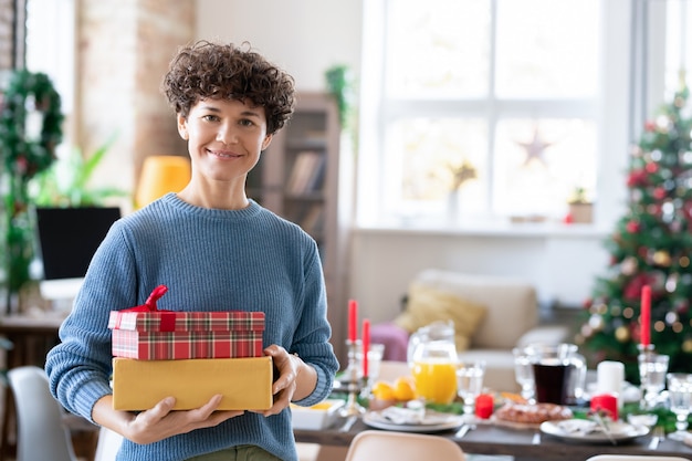 Giovane donna bruna riccia sorridente che tiene scatole regalo con regali per la sua famiglia mentre si trova contro l'albero di Natale decorato e il tavolo servito