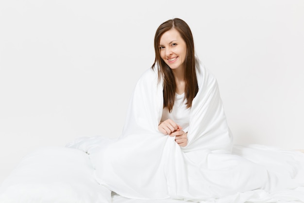 Giovane donna bruna seduta a letto con lenzuolo bianco, cuscino, avvolto in una coperta sul muro bianco