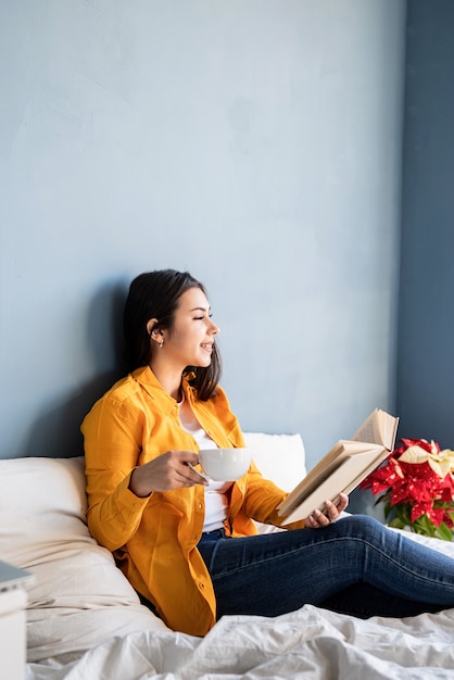giovane donna bruna seduta nel letto con mangiare croissant e leggere un libro