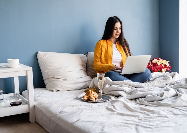 Giovane donna bruna seduta nel letto lavorando sul computer bevendo champagne