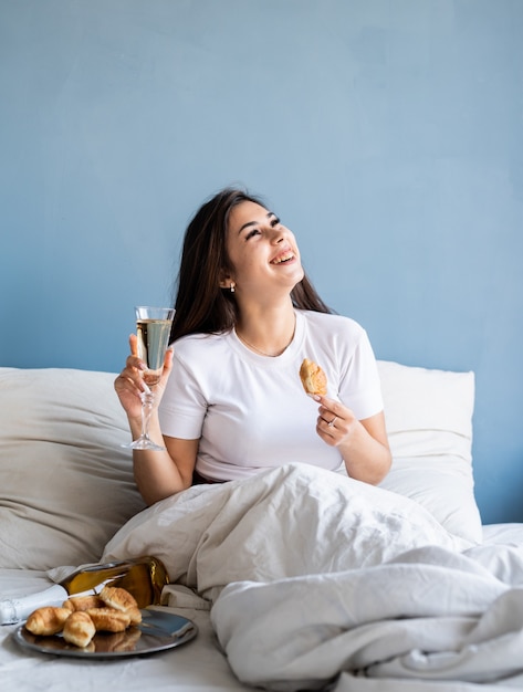 Giovane donna bruna seduta sveglia nel letto bevendo champagne mangiando croissant