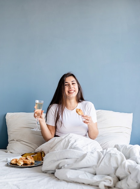 giovane donna bruna seduta sveglia nel letto bevendo champagne mangiando croissant