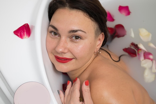 Giovane donna bruna sorridente che fa un bagno con petali di rosa a casa facendo trattamenti termali per l'equilibrio e la salute mentale