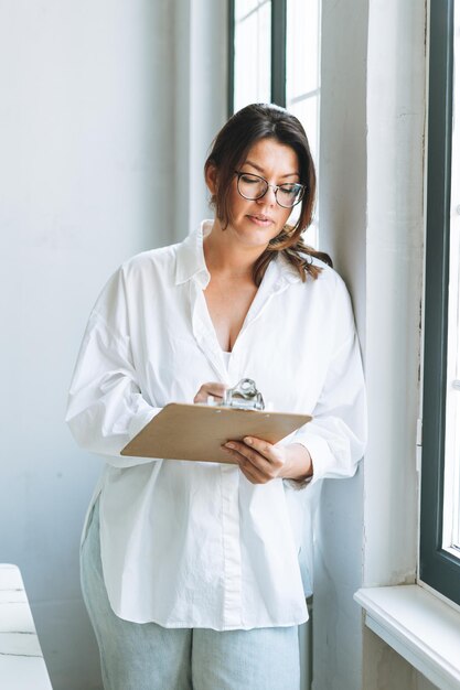 Giovane donna bruna sorridente plus size in camicia bianca vicino alla finestra con penna e documenti in mano nell'ufficio moderno e luminoso