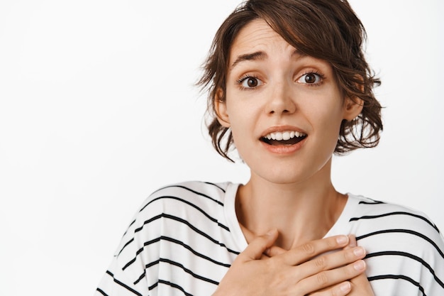 Giovane donna bruna sospirando, tenendo le mani sul petto e guardando delicatamente la fotocamera, sfondo bianco.