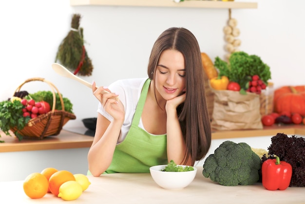 Giovane donna bruna sta cucinando o mangiando insalata fresca in cucina. Casalinga che tiene un cucchiaio di legno nella mano destra. Concetto di cibo e salute.