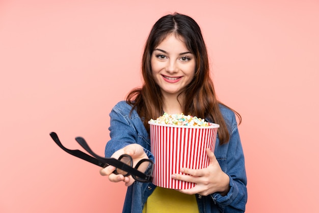 Giovane donna bruna sul muro rosa con gli occhiali 3d e in possesso di un grande secchio di popcorn