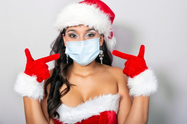 Giovane donna bruna vestita con il costume di babbo natale che le indica la sua maschera di protezione antivirus