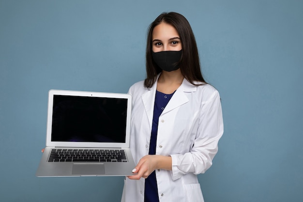 Giovane donna brunetta che indossa camice bianco medico e maschera nera che tiene in mano un computer portatile e guarda la telecamera isolata su sfondo blu muro