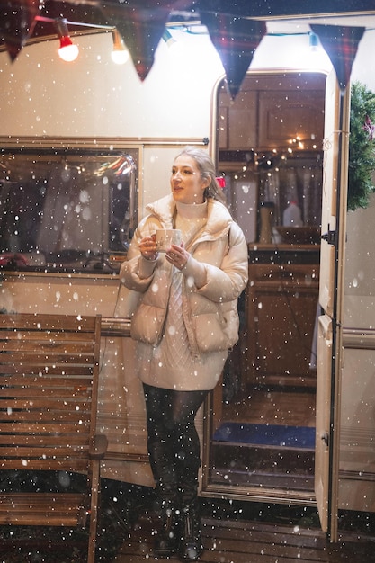 Giovane donna candida in piena altezza godimento rilassante tazza di caffè o tè in mano che tiene felice anno nuovo e notte di Natale alla porta nel rimorchio mobile di viaggio all'aperto. neve che cade