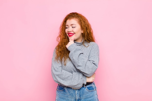 Giovane donna capa rossa che sorride con un'espressione felice e sicura con la mano sul mento