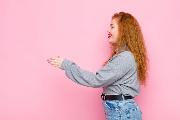 Giovane donna capa rossa che sorride, ti accoglie e offre una stretta di mano per chiudere un affare riuscito, concetto di cooperazione sopra la parete rosa