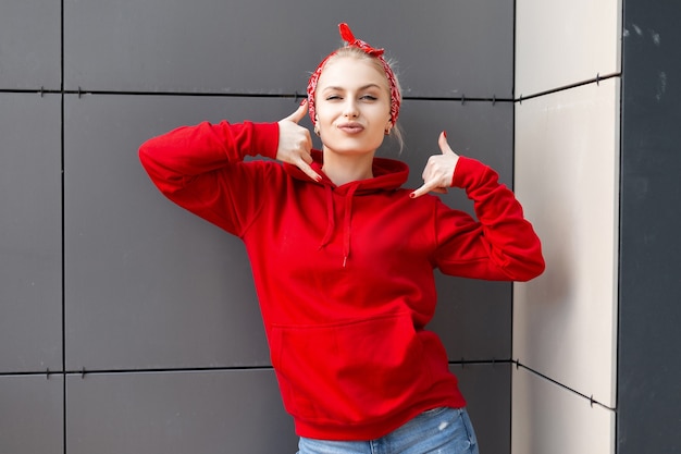 Giovane donna carina divertente in felpa rossa vintage con cappuccio con una bandana alla moda in jeans pone in città vicino a un edificio grigio in una giornata estiva