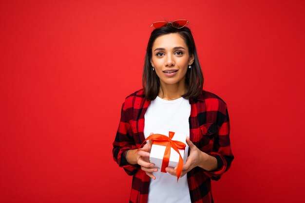 Giovane donna castana abbastanza positiva isolata sopra il muro rosso che indossa la maglietta casuale bianca e la camicia rossa e nera che tiene il contenitore di regalo bianco con il nastro rosso