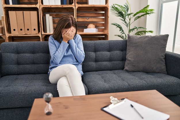 Giovane donna castana all'ufficio di consultazione con espressione triste che copre il viso con le mani mentre piange. concetto di depressione.