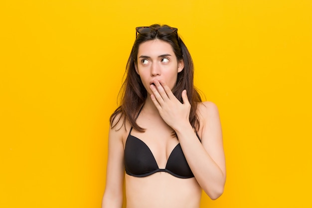 Giovane donna castana che indossa un bikini contro la parete gialla che sbadiglia mostrando un gesto stanco che copre la bocca con la mano.