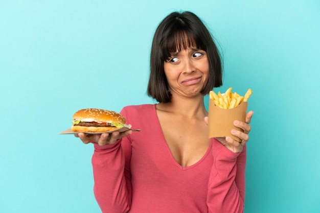 Giovane donna castana che tiene hamburger e patatine fritte sopra la superficie blu isolata