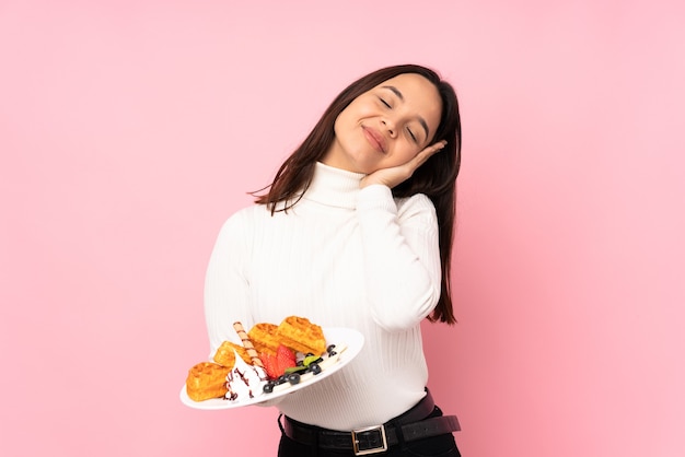 Giovane donna castana che tiene i waffle sopra fondo rosa isolato che fa gesto di sonno nell'espressione dorable