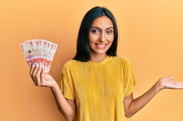Giovane donna castana che tiene in mano 10 pesos colombiani banconote che celebrano il successo con un sorriso felice e l'espressione del vincitore con la mano alzata
