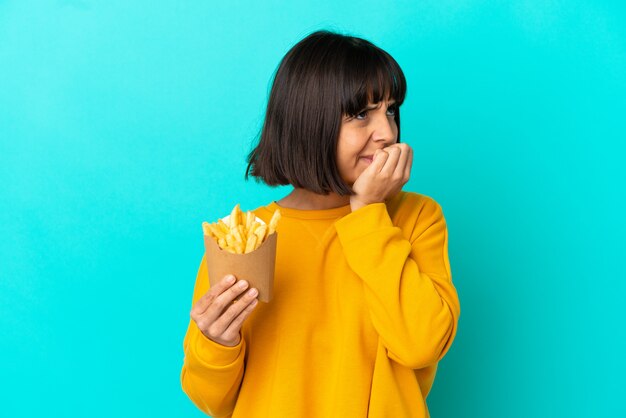 Giovane donna castana che tiene le patatine fritte sopra la superficie blu isolata che ha dubbi?