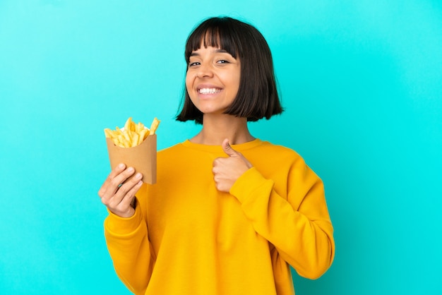 Giovane donna castana che tiene patatine fritte su sfondo blu isolato dando un gesto di pollice in alto