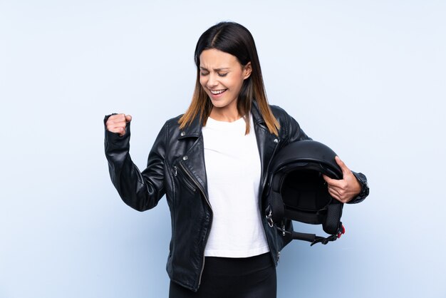 Giovane donna castana che tiene un casco del motociclo sopra la parete blu che celebra una vittoria