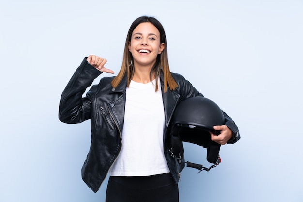 Giovane donna castana che tiene un casco del motociclo sopra la parete blu fiera e soddisfatta di sé