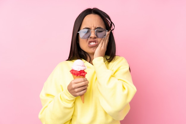 Giovane donna castana che tiene un gelato della cornetta sopra la parete rosa isolata con mal di denti