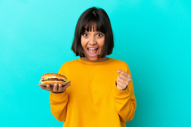 Giovane donna castana che tiene un hamburger sopra fondo isolato che celebra una vittoria nella posizione del vincitore