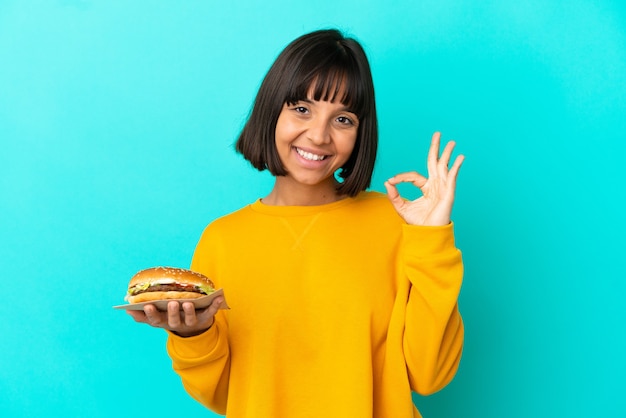 Giovane donna castana che tiene un hamburger sopra fondo isolato che mostra il segno giusto con le dita