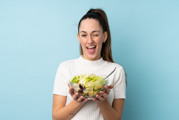 Giovane donna castana che tiene un'insalata sopra la parete blu isolata