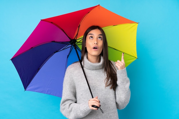 Giovane donna castana che tiene un ombrello sopra la parete blu isolata che indica con il dito indice una grande idea