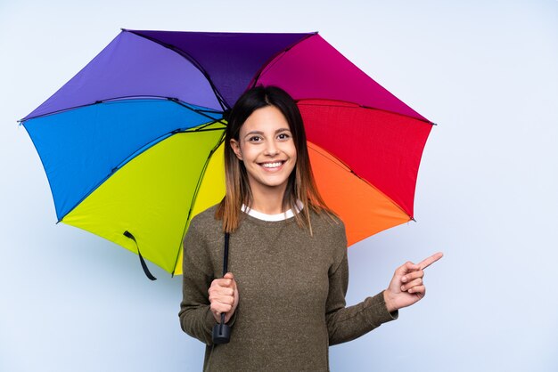 Giovane donna castana che tiene un ombrello sopra la parete blu isolata che indica dito il lato