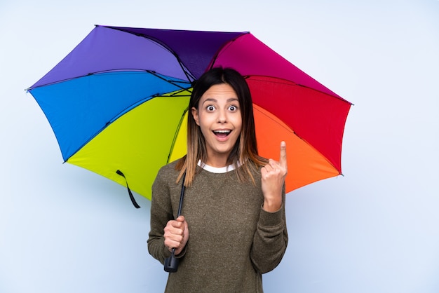 Giovane donna castana che tiene un ombrello sopra la parete blu isolata con espressione facciale di sorpresa