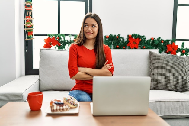 Giovane donna castana che usa il computer portatile seduto sul divano a Natale sorridendo guardando di lato e fissando lontano pensando